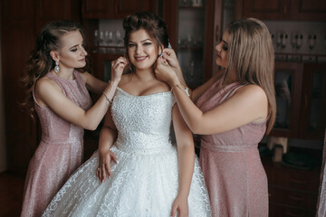 Three women are getting ready for a wedding. One of them is putting on a wedding dress. The other two women are helping her with her hair and makeup