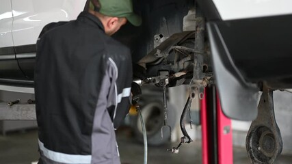 A mechanic in a uniform is shown from the waist down repairing the underside of a white car that is mounted on a hydraulic lift.