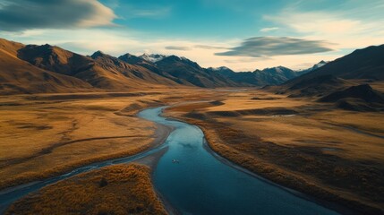 Fototapeta premium A serene river gracefully flows through vibrant golden valleys, surrounded by towering mountains that touch the clear blue sky, capturing nature's tranquil beauty at dusk