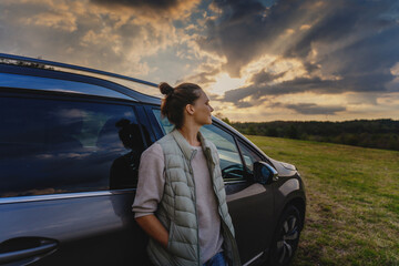 Young female driver standing by car enjoying sunset and nature in field during road trip