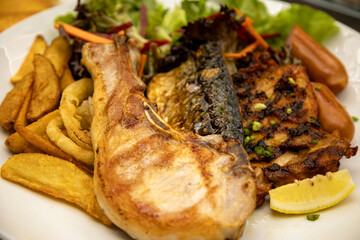 Grilled pork chop with crispy potato wedges, grilled fish fillet, fresh salad, sausages, and lemon wedge on white plate