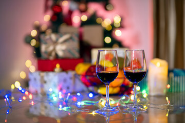 Two wine glasses filled with red wine placed on table with colorful fairy lights, in festive with blurry holiday decorations and gift boxes in background