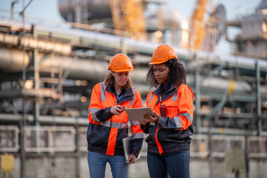 Engineers wearing safety gear, including hard hats examining survey a large blueprint tablet standing industrial facility gas or oil refinery engaged in a job requires high safety standards concept