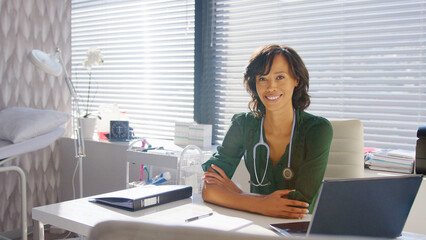 Portrait Of Smiling Female Doctor With Stethoscope Sitting Behind Desk In Office Or Surgery