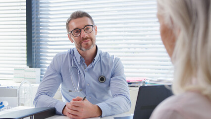 Mature Female Patient Meeting With Doctor Sitting At Desk In Office Or Surgery