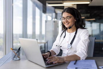 Confident female doctor wearing glasses and headset engaged in online consultation using laptop. Stethoscope around neck signifies professional medical setting.