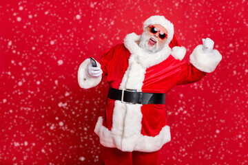 Portrait of crazy santa claus in hat using cell phone device raising fists screaming yes having newyear party wearing belt eyewear eyeglasses isolated over red background