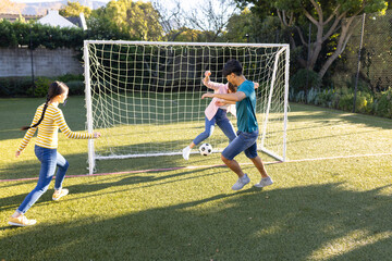 Playing soccer, children kicking ball towards goal on sunny day in backyard