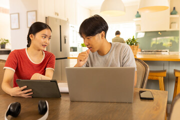 Collaborating on project, young asian brother and sister using tablet and laptop in kitchen