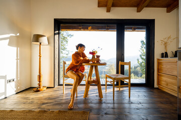 A woman in a warm, oversized sweater sits at a wooden table, gazing out at the serene landscape...