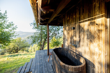 Rustic outdoor wooden bathtub on a cabin deck, overlooking the lush green landscape and hills, offering a peaceful escape in nature