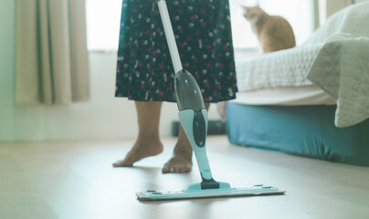 A woman is cleaning the floor with a mop. A cat is sitting on the bed