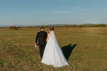 A bride and groom are walking in a field. The bride is wearing a white dress and the groom is wearing a black suit. The couple is holding hands and looking at each other. The field is empty