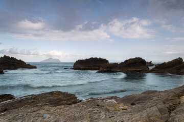 At dusk. Gentle waves lap against smooth, rounded rocks in a tranquil cove. The sky is painted in soft hues of pink and purple, reflecting on the calm water. Toucheng, Yilan, Taiwan.