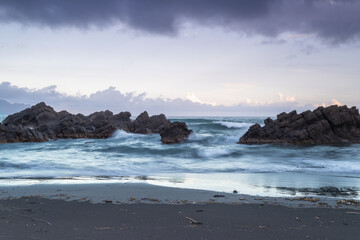 At dusk. Gentle waves lap against smooth, rounded rocks in a tranquil cove. The sky is painted in soft hues of pink and purple, reflecting on the calm water. Toucheng, Yilan, Taiwan.