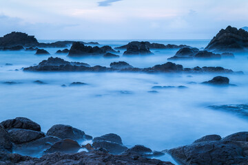 At dusk. Gentle waves lap against smooth, rounded rocks in a tranquil cove. The sky is painted in soft hues of pink and purple, reflecting on the calm water. Toucheng, Yilan, Taiwan.