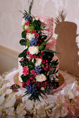 A tall cake with flowers on it. The cake is pink and white. The flowers are in a long line and are of different colors