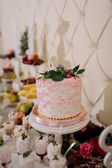 A white cake with pink frosting and flowers on top sits on a white cake stand. The cake is surrounded by a variety of fruit and dessert items, including a bowl of strawberries and a plate of bananas