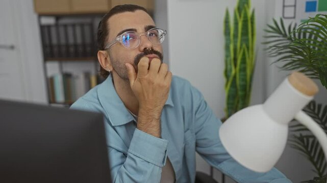 Handsome man with moustache in office deep in thought wearing glasses and blue shirt at desk surrounded by plants
