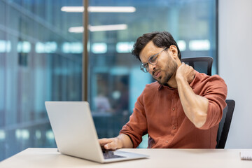 Man using laptop in office shows neck pain and discomfort, representing challenges related to...