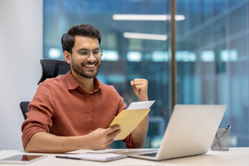 Fototapeta premium Smiling man in office holds envelope, reads positive news with excitement. Seated at desk with laptop . Success and happiness expressed through raised fist, embodying achievement and motivation.