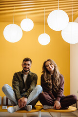 Beautiful young couple having fun in the room. A man and a woman are sitting and smiling against the background of an orange wall and large round lamps. Portrait of a boy and a girl looking at camera.