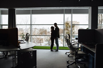 Men and women flirting in the workplace. Silhouette of a man and a woman hugging and standing near a panoramic window. Romantic kisses in office. Beautiful young colleagues have a relationship at work
