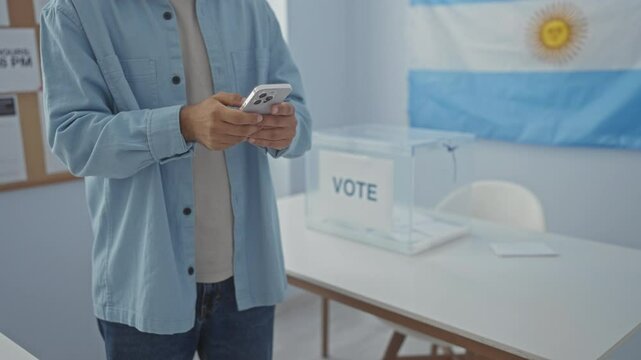 Hispanic man using smartphone in an electoral college room with an argentinian flag, banner, and a transparent ballot box on the table.