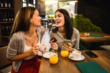 Two girl best friends met at the cafe to shop online together. They are using mobile phone and credit card. Friends hang out and drink coffee and orange juice.