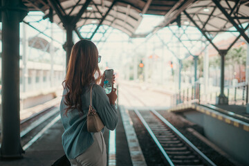 A woman stands at a busy train station, focused on her smartphone, waiting for her train