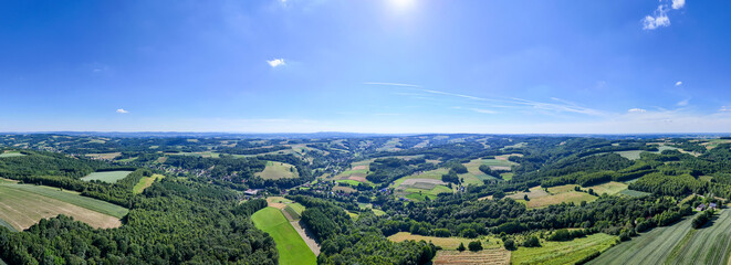 Aerial panorama of a lush green landscape, rolling hills, forests, and farmlands under a bright blue sky with scattered clouds, captured on a sunny day