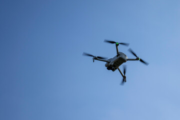 White drone flying in the sky against a clear blue background. The drone features green lights on its propellers. Ideal for technology and aerial photography themes.
