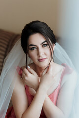 A woman in a pink dress is posing for a photo with her hands clasped behind her back. She has a veil in her hair and is wearing a necklace. The photo has a romantic and elegant mood