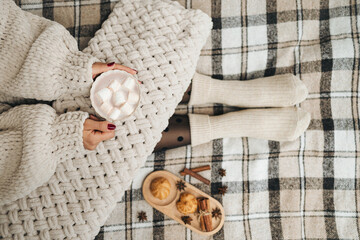 Woman in a knitted sweater sitting on a cozy plaid blanket, holding a cup of hot chocolate or cocoa drink with marshmallows. Hygge concept