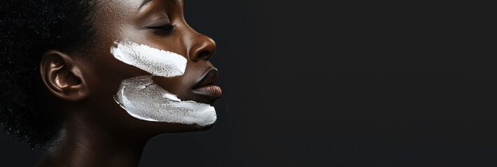 African American woman close-up face applies white cream in beauty studio. She focuses on her skin tone, showcasing makeup skills. Dark background enhances facial features and product application.