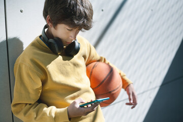 A boy in a yellow sweater holds a basketball and looks at his mobile phone.