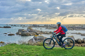 Obraz premium nice senior woman cycling with her electric mountain bike at the rocky rose granite coast at Meneham site in Brittany, France next to Kerouarn