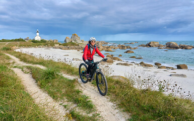 nice senior woman cycling with her electric mountain bike at the rocky rose granite coast  at Meneham site in Brittany, France next to Kerouarn