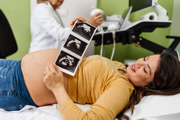 Beautiful and pregnant woman is happy after pregnancy medical check up. She is holding and looking at ultrasound scan of her baby. Modern pregnancy healthcare concept.