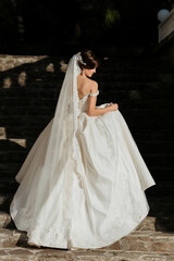 Naklejka premium A woman in a white wedding dress is walking down a set of stairs. The dress is long and has a lace-like design. The woman is holding her dress up to her face, possibly adjusting it or admiring it