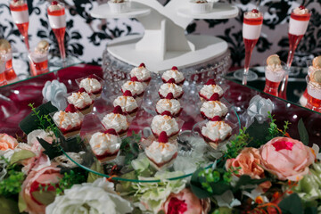 A table with a variety of desserts, including cupcakes and cakes, and a floral arrangement. The desserts are displayed on a glass plate, and there are several wine glasses and bottles nearby