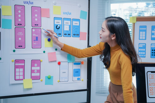 Ux ui designer is reviewing mobile phone application wireframes displayed on a board in the office. She is using a pen to point at specific elements of the design