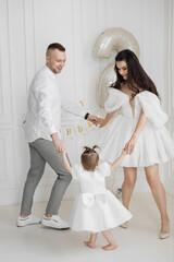 Obraz premium Caucasian family celebrating child's birthday. Parents dancing with young daughter in white room decorated with happy birthday banner. Father mother and child dressed elegantly sharing joyful moment