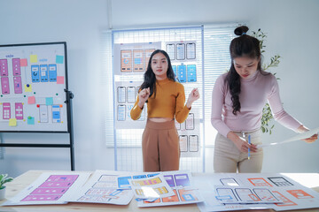 Two asian female designers collaborate on a mobile app wireframe in a vibrant office setting. They...