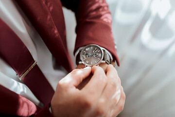 A man in a red suit is wearing a watch and adjusting it. The watch is silver and has a black face. The man is wearing a tie and a jacket, and he is in a professional setting