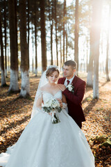 A bride and groom are standing in a forest, with the bride wearing a white dress and the groom wearing a red suit