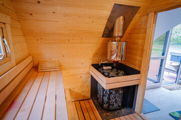 A cozy wooden sauna interior with benches and a traditional sauna stove, featuring hot stones and a stainless steel water heater for steam..