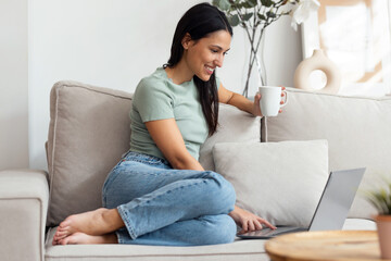 Pretty young woman working with her laptop while drinking a cup of coffee sitting on a couch at home