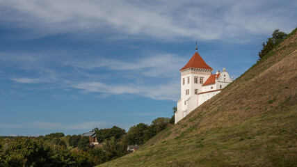 Fototapeta premium white tower with red roof of old grodno castle behind green grassy slope of big hill under blue sky on fine september day. side view with copy space