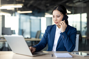 Asian businesswoman wearing blue suit engaged in multitasking at work. Using laptop and phone in...